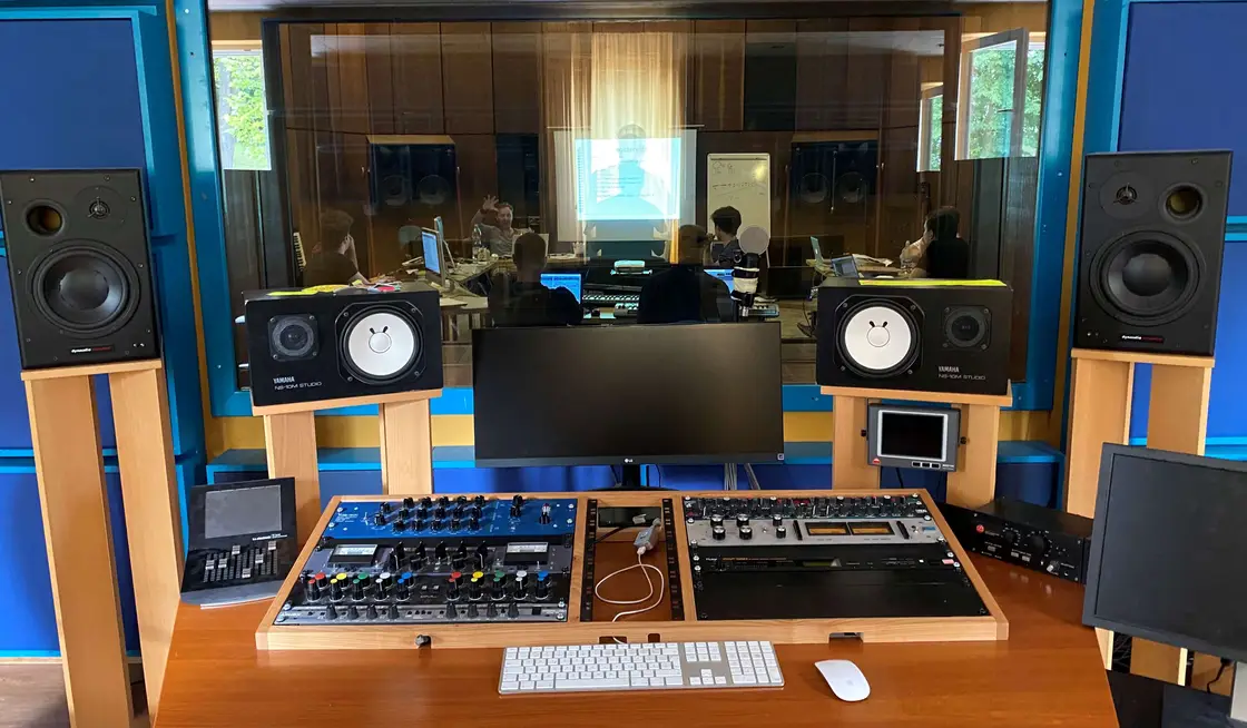 Studio control room: Yamaha NS-10M and Dynaudio monitors on wooden stands, analogue outboard mounted in the desk, and a seminar visible through the glass into the live room.