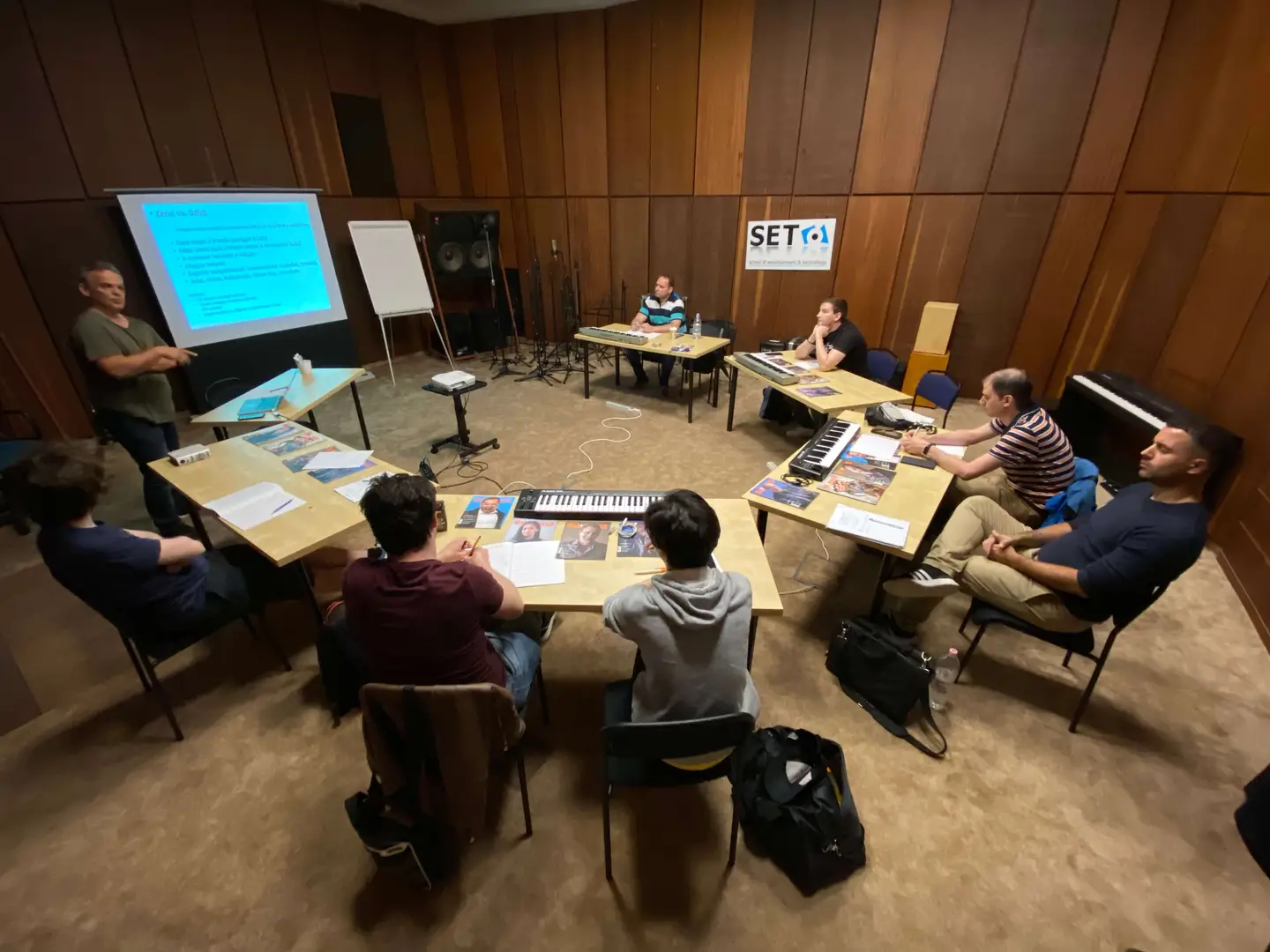 A seminar in progress in the wood-panelled Budapest room: participants sit at tables with MIDI keyboards and printed material while a teacher presents at the projection screen.