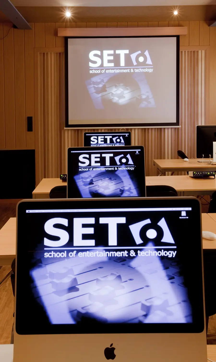 Workstation room with a Mac and an audio interface on every desk, the SET School logo shown on each screen and on the projection screen behind them.